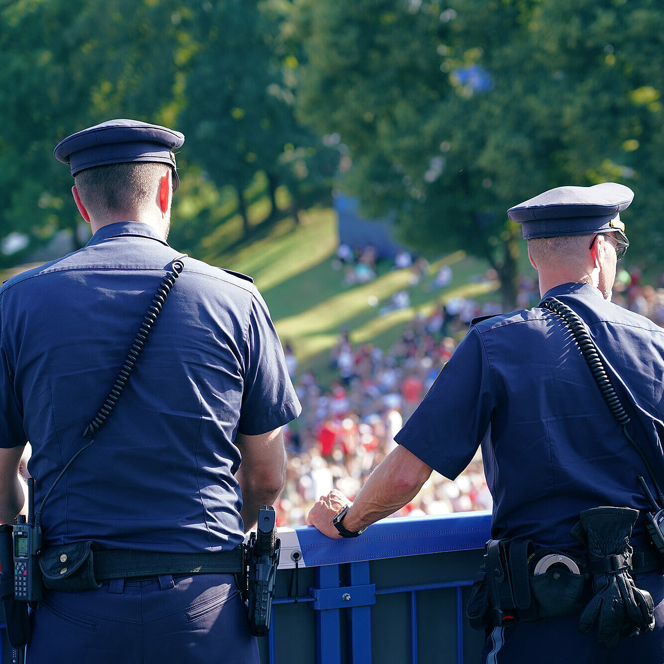 Zwei Polizeibeamte in Uniform stehen mit dem Rücken zur Kamera und beobachten eine im Freien versammelte Menschenmenge.