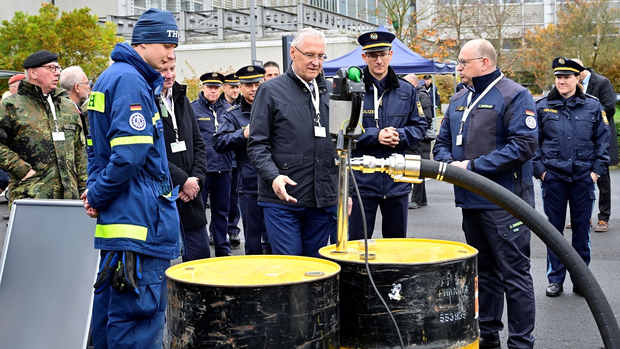Auf dem Bild ist Innenminister Joachim Herrmann gemeinsam mit Polizisten und Einsatzkräften von der Bundeswehr und dem Technischen Hilfswerk zu sehen, wie diese gemeinsam an Übungsmaterial der MainTEX stehen.