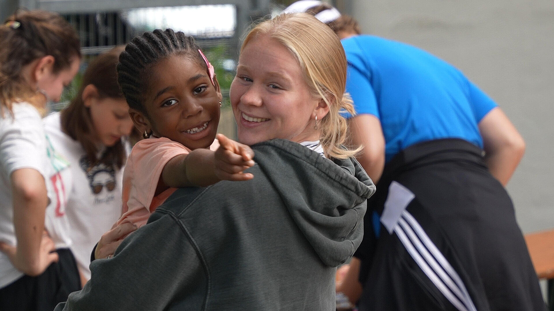 Mehrere Kinder stehen zusammen im Freien, einige mit Händen auf den Hüften, während eine Person mit blonder Haarfarbe und grauem Hoodie in den Vordergrund schaut. Die Kinder tragen unterschiedliche Kleidung, und im Hintergrund sind weitere Kinder zu sehen.