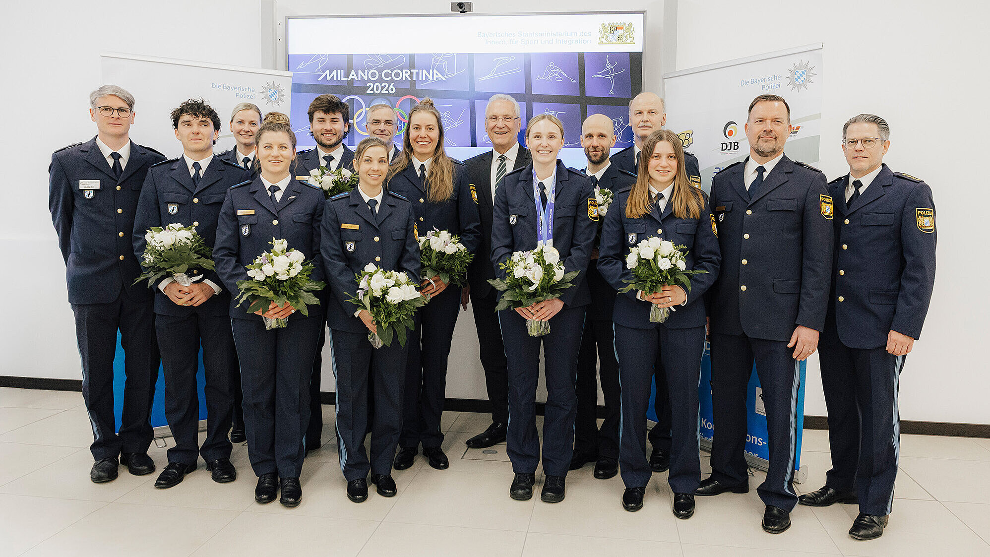 Gruppenfoto mehrere Personen in dunkelblauen Polizeiuniformen, einige mit weißen Blumensträußen, vor einer Leinwand mit „MILANO CORTINA 2026“