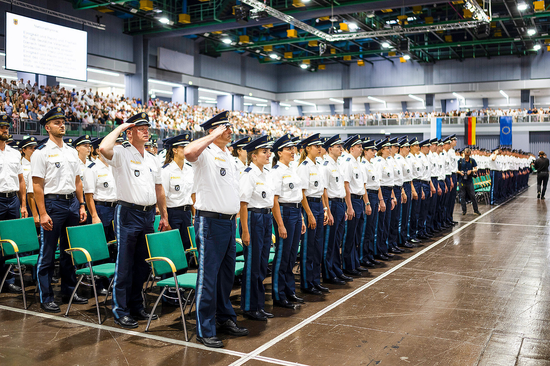 Viele junge Polizistinnen und Polizisten bei Vereidigungszeremonie der Bayerischen Polizei in einer großen Halle, einige salutieren