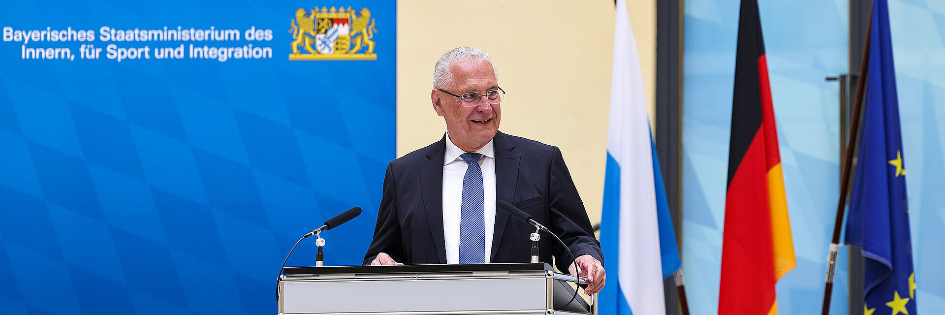 Innenminister Herrmann lächelnd am Rednerpult, im Hintergrund Fahnen und Pressewand des StMI