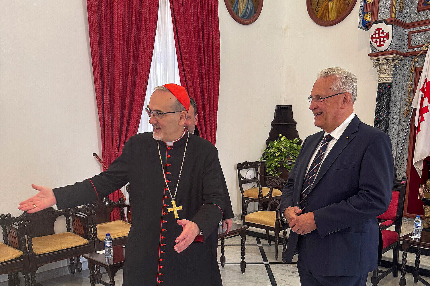 Kardinal Pizzaballa in traditioneller schwarzer Robe mit rotem Besatz und Kreuzkette begrüßt freundlich Innenminister Joachim Herrmann in einem festlich geschmückten Raum mit roten Vorhängen, Ikonen und kunstvoller Wandgestaltung.“