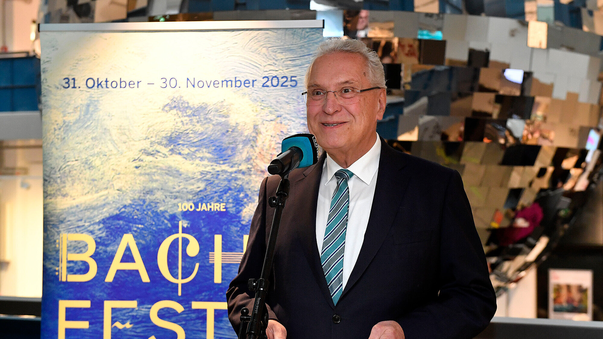  beschreibt eine Person in formellInnenminister Joachim Herrmann mit Blick in die Kamera. Im Hintergrund ist ein Plakat zu sehen, das das Bachfest München 2025 ankündigt, das vom 31. Oktober bis zum 30. November 2025 stattfindet.