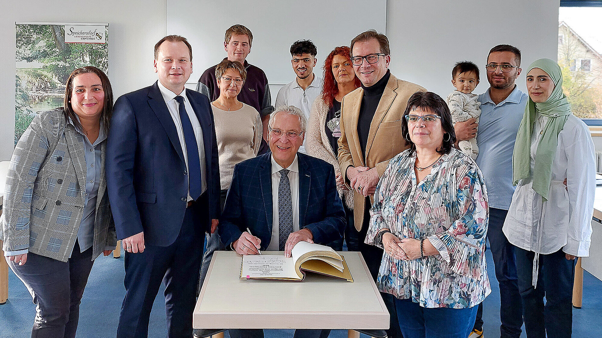 Eine Gruppe von Personen, am Tisch sitzend Innenminister Joachim Herrmann, zeigt sich lächelnd vor dem goldenen Buch der Stadt in einem Foto.