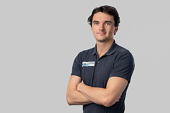 Vicktor Noah vor hellem Hintergrund, trägt dunkles Poloshirt mit rechteckigem Aufnäher und hat die Arme verschränkt.