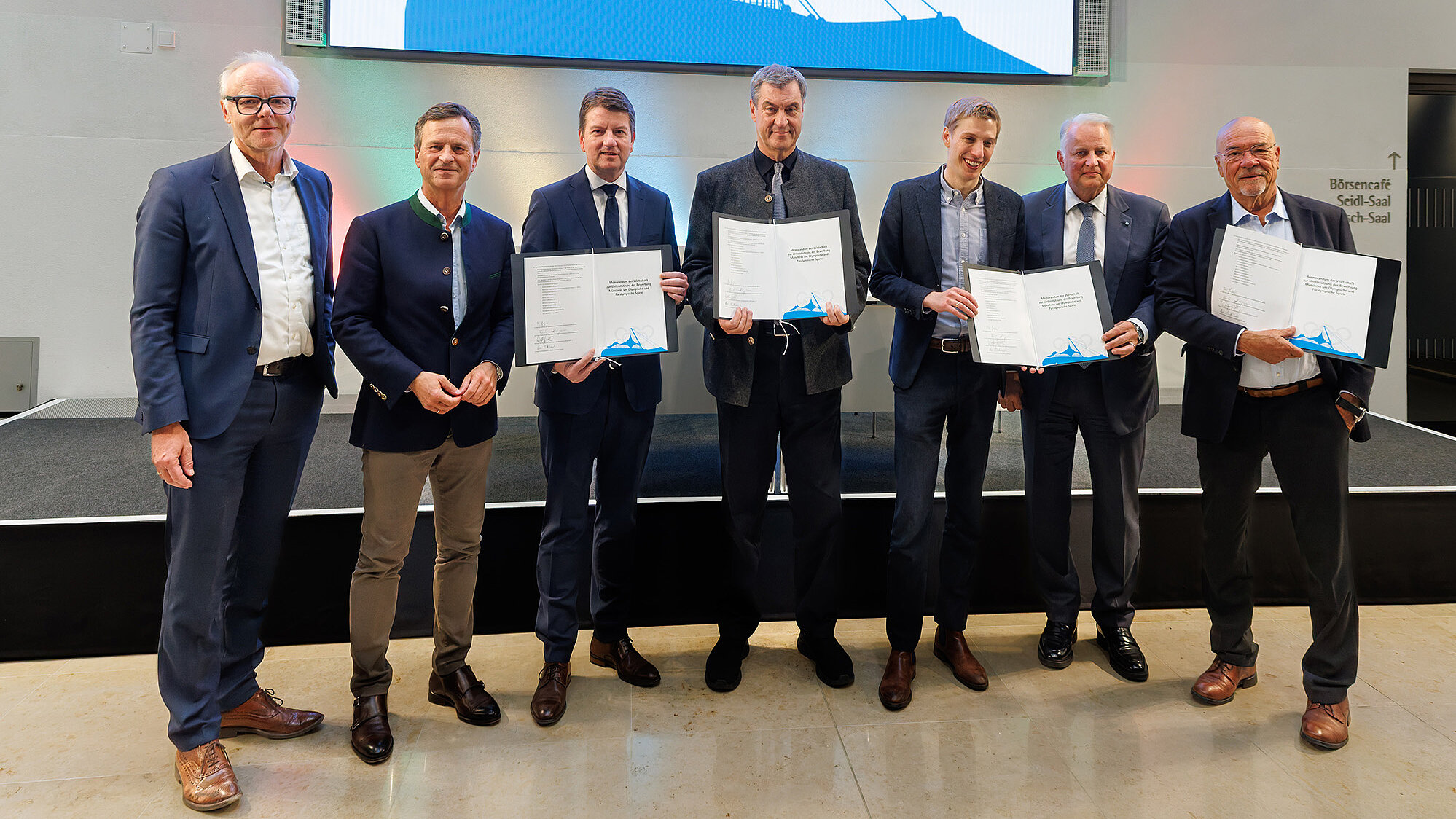 Sieben Männer in Anzügen und Jacken stehen nebeneinander vor einer Bühne und halten Urkunden; im Hintergrund ein großes Display mit blauem Grafikelement.