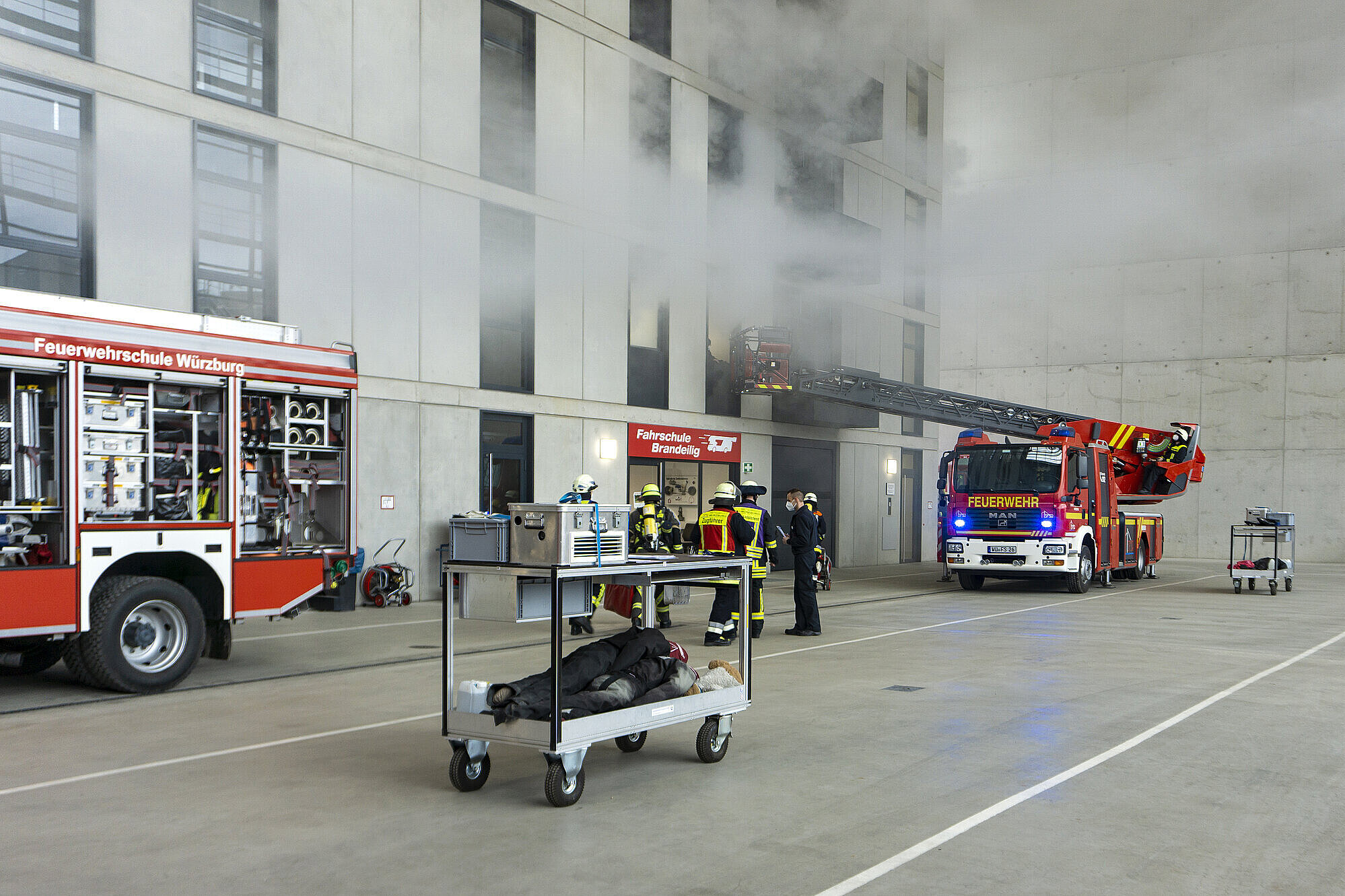 Übung der Feuerwehrschule bei der mehrere Feuerwehrautos und Feuerwehrmänner bei Balkons an einer Häuserfassade stehen, aus denen Rauch austritt