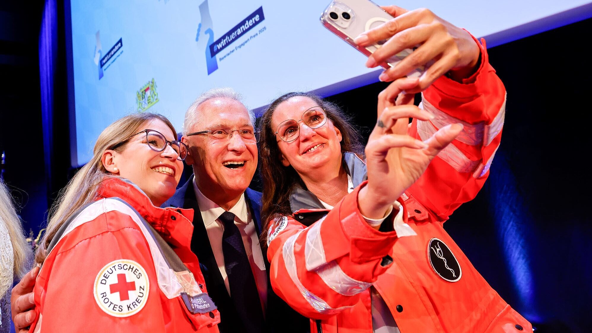 Auf dem Bild ist Innenminister Joachim Herrmann zu sehen, wie er mit zwei BRK-Mitarbeiterinnen ein Selfie macht.