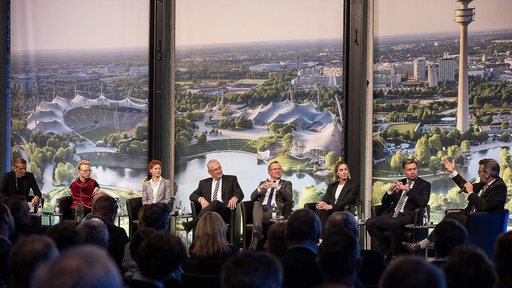 Podiumsdiskussion mit sieben Personen auf einer Bühne vor einem großen Panoramafenster, das einen Blick auf den Olympiapark München mit dem Olympiastadion, Zeltdachbauten und dem Olympiaturm zeigt. Im Vordergrund ist ein Publikum zu sehen.