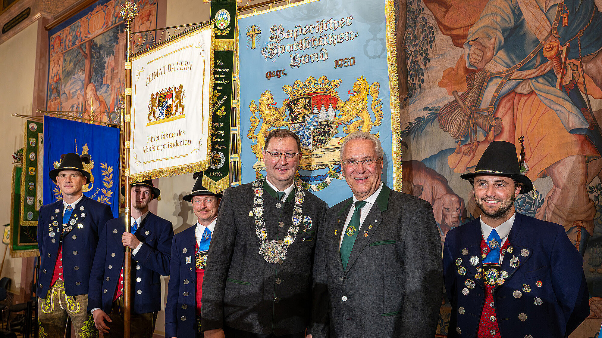 Innen- und Sportminister Joachim Herrmann (rechts) mit dem Präsidenten des Bayerischen Sportschützenbundes, Christian Kühn (Mitte), und weiteren festlich gekleideten Schützen bei der Jubiläumsfeier zum 75-jährigen Bestehen des BSSB. Im Hintergrund sind prächtige Fahnen und Wandteppiche zu sehen.