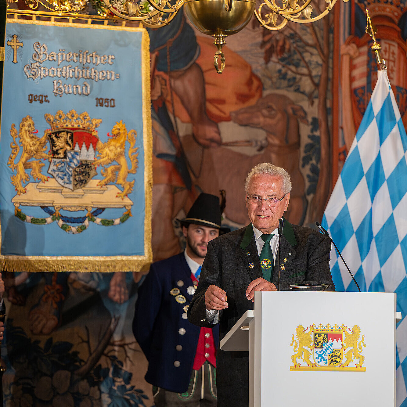 Bayerns Innen- und Sportminister Joachim Herrmann  bei der Jubiläumsfeier zum 75-jährigen Bestehen des BSSB am Rednerpult. Im Hintergrund sind prächtige Fahnen und Wandteppiche zu sehen.