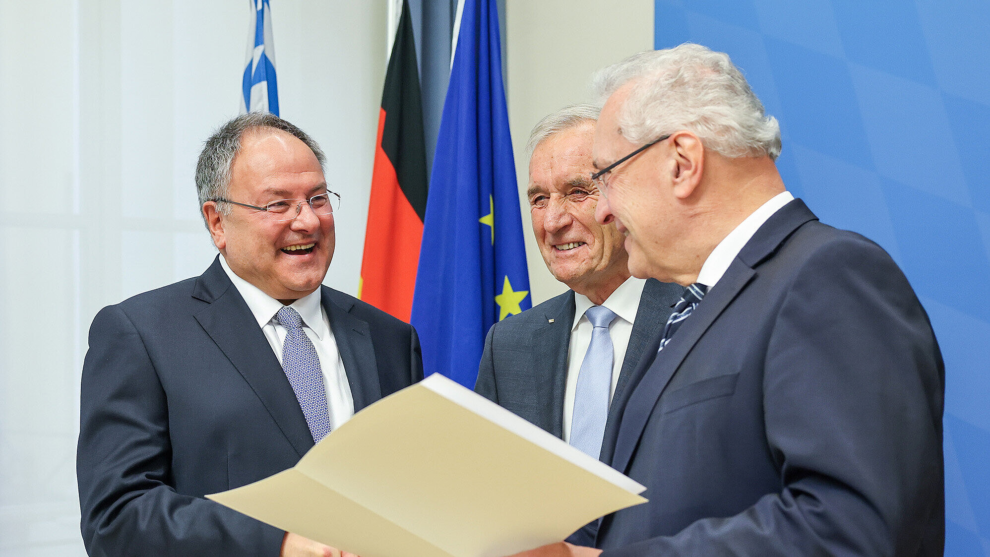 Drei Männer (1. von rechts Innenminister Joachim Herrmann) in Anzügen stehen in einem Raum mit blauer Wand und bayerischer sowie europäischer Flagge im Hintergrund. Zwei von ihnen geben sich die Hand, während der dritte Mann ein geöffnetes Dokument hält und es zeigt. Die Szene wirkt formell und erinnert an eine Preisverleihung oder offizielle Übergabe.