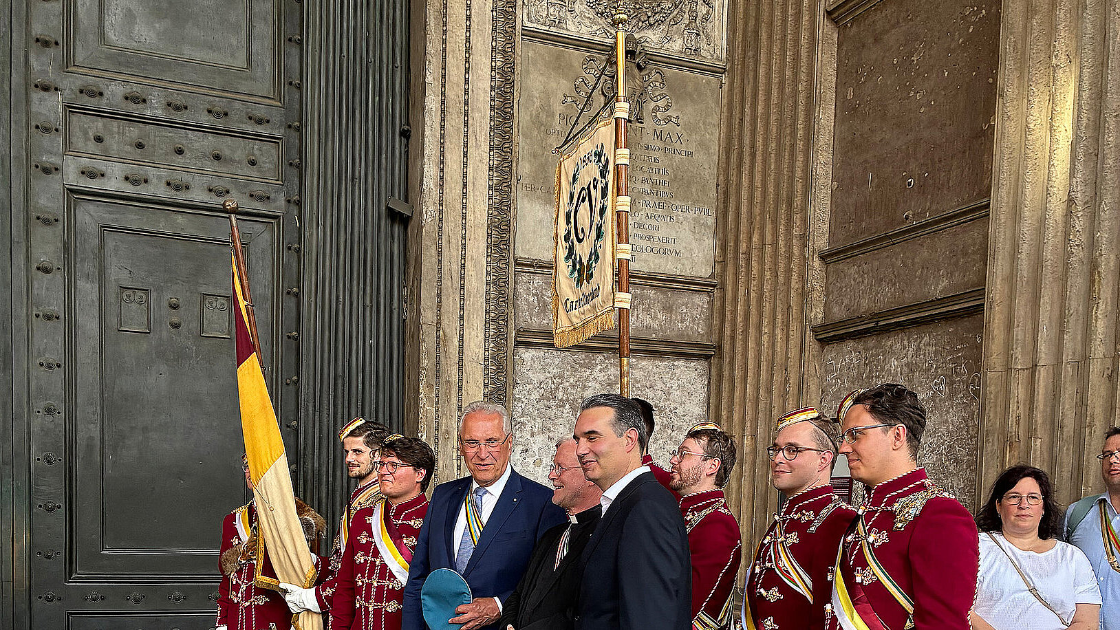 Eine Gruppe von Personen, teils in festlicher, traditioneller Kleidung, steht vor einem großen, verzierten Tor. Einige tragen rote Uniformen mit goldenen Details und Stiefeln, während andere in Anzügen gekleidet sind. Im Hintergrund ist ein Banner mit einem Wappen zu sehen.
