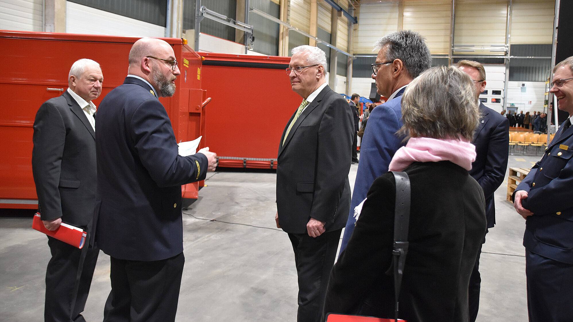 Mehrere Personen — Männer in Anzügen und einer Uniform sowie eine Frau mit pinkem Schal — stehen in einer großen Industriehalle vor großen roten Containern. Zwei Personen führen ein Gespräch, im Hintergrund sind Stuhlreihen und weitere Anwesende erkennbar.