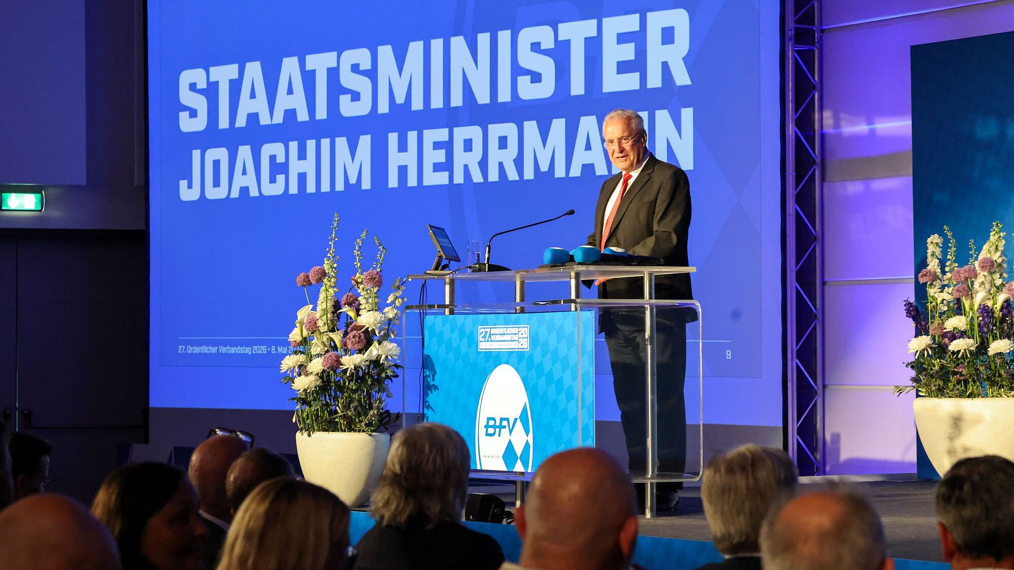 Innenminister Herrmann am Rednerpult auf einer Bühne, hinter ihm eine Präsentation auf dem sein Name steht, vor ihm aufmerksames Publikum.