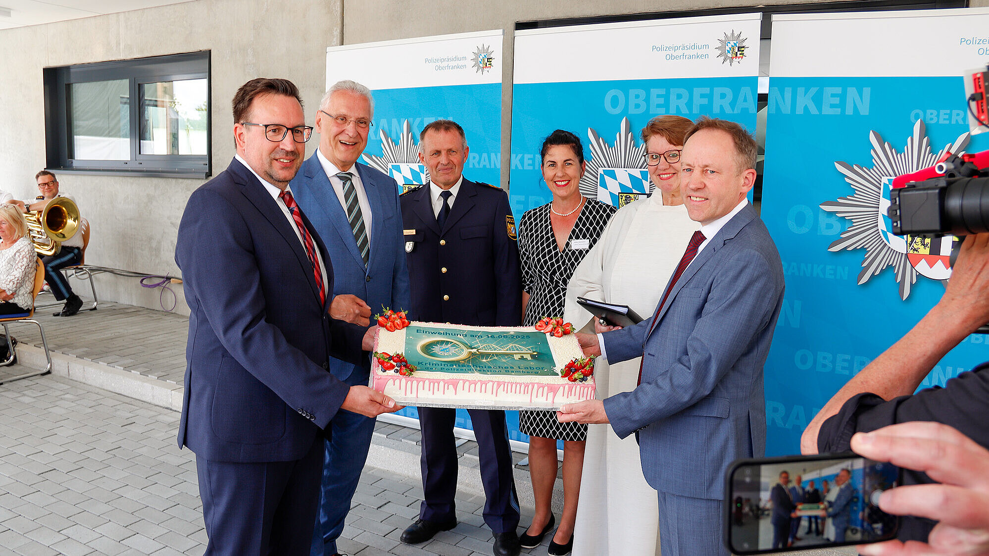  Mehrere festlich gekleidete Personen stehen vor zwei Roll-Ups mit dem Logo des Polizeipräsidiums Oberfranken. Zwei Männer im Anzug halten eine dekorative Torte mit der Aufschrift „Einweihung 14.06.2024 – Kriminallabor Bamberg“. Im Hintergrund sind eine Polizistin, ein Polizeibeamter, eine Geistliche sowie eine Musikerin mit Tuba zu sehen. Eine Person filmt die Szene mit dem Smartphone. Feierlicher Anlass in einem modernen, offenen Gebäudebereich.