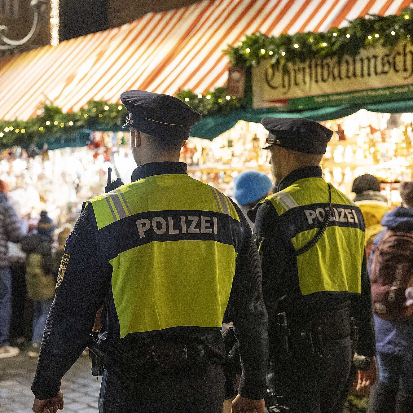 Zwei Polizisten in gelben Warnwesten mit der Aufschrift „POLIZEI“ gehen bei Nacht über einen belebten Christkindlesmarkt in Nürnberg, im Hintergrund sind Marktstände mit Weihnachtsdekoration und mehrere Besucher zu sehen.