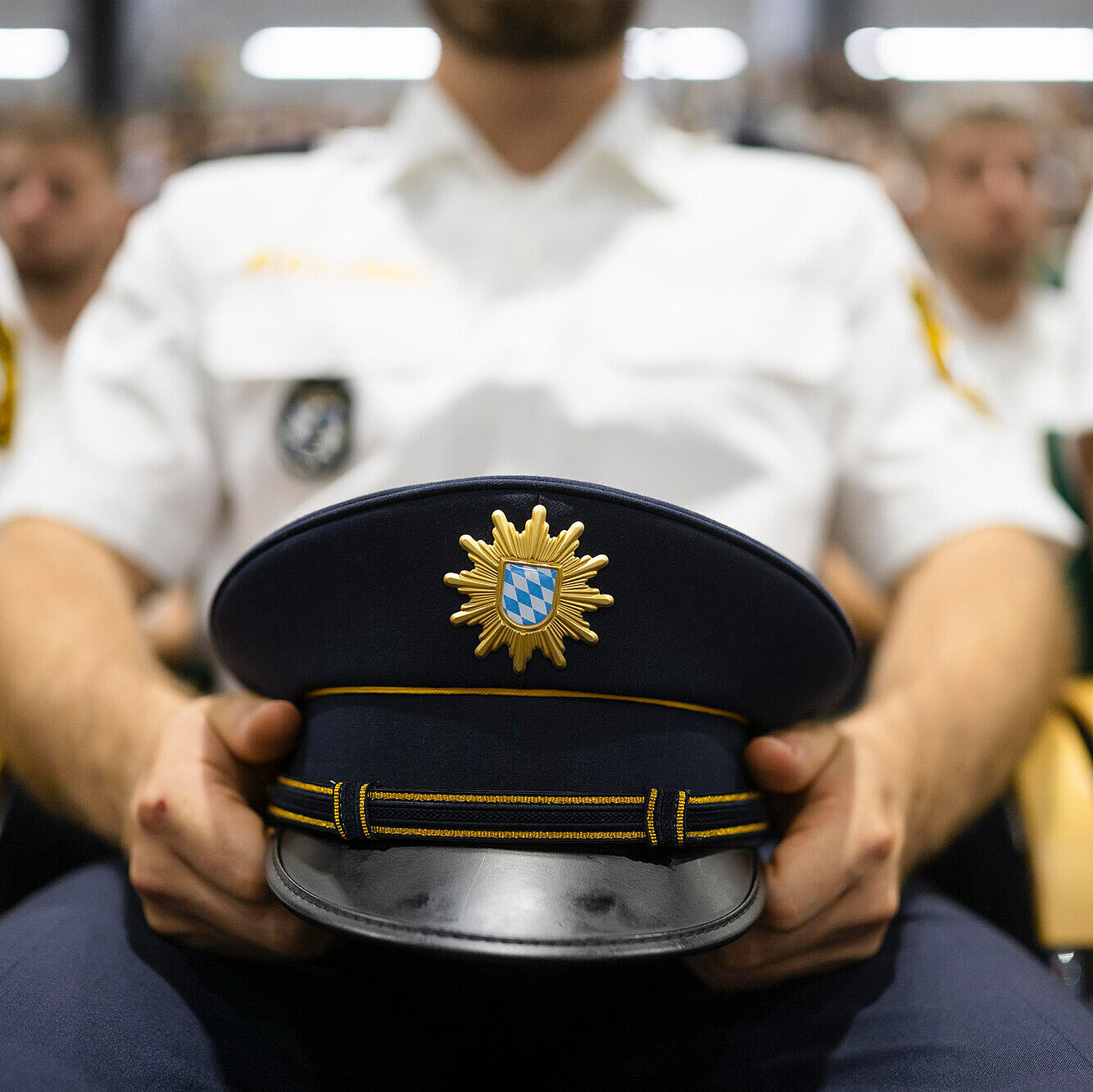 Polizist sitzend in weißem Polizeihemd hält seine Polizeimütze mit Sympbol bayerische Polizei auf dem Schoß