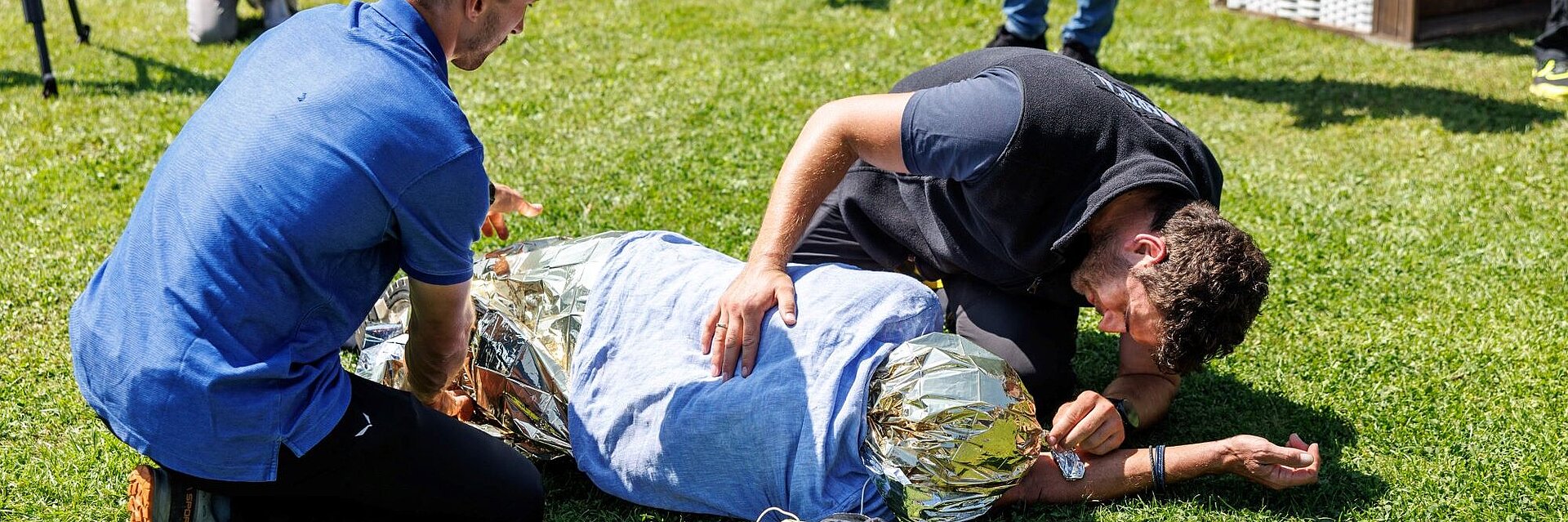 Zwei Personen leisten Erste Hilfe bei einer verletzten Person, die auf einer Wiese liegt und mit einer Decke zugedeckt ist