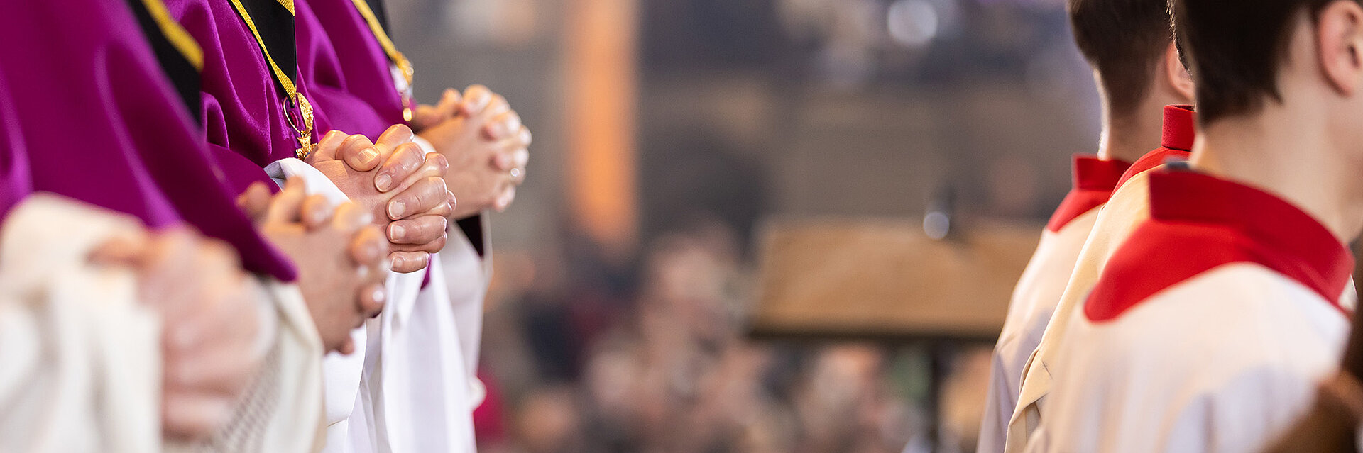 Nahaufnahme von liturgisch gekleideten Personen mit gefalteten Händen in einer Kirchenbank; unscharfer Innenraum und Gemeinde im Hintergrund.