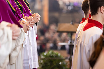 Nahaufnahme von liturgisch gekleideten Personen mit gefalteten Händen in einer Kirchenbank; unscharfer Innenraum und Gemeinde im Hintergrund.