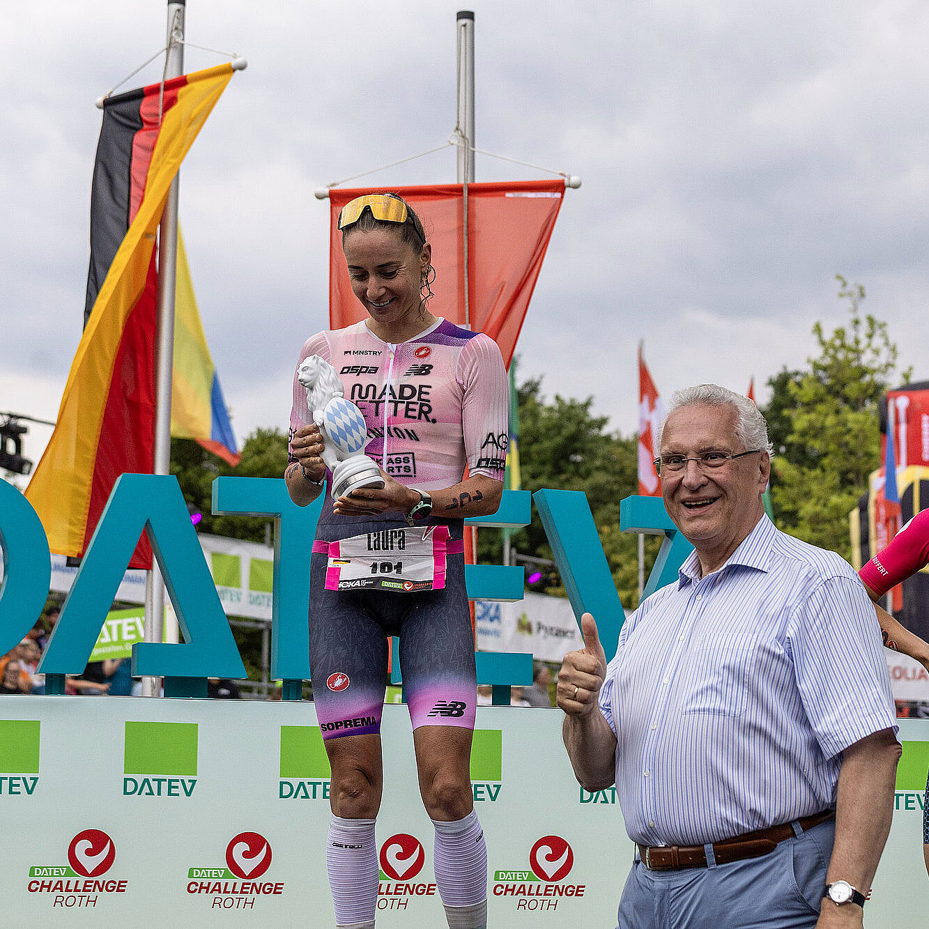 Sportminister Herrmann neben Siegerin, Laura Philipp, die auf Siegertreppchen steht, im Hintergrund bunte Fahnen und das Logo DATEV