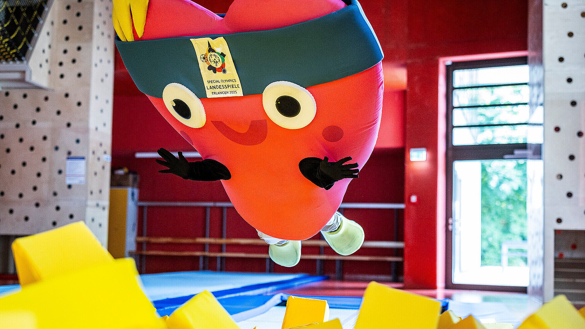 Ein großes, rotes Maskottchen in Herzform mit Stirnband und breitem Lächeln springt in eine gelbe Schaumstoffgrube in einer Sporthalle. Auf dem Stirnband ist ein Schild mit dem Logo und der Aufschrift „Special Olympics Landesspiele Erlangen 2025“ zu sehen.