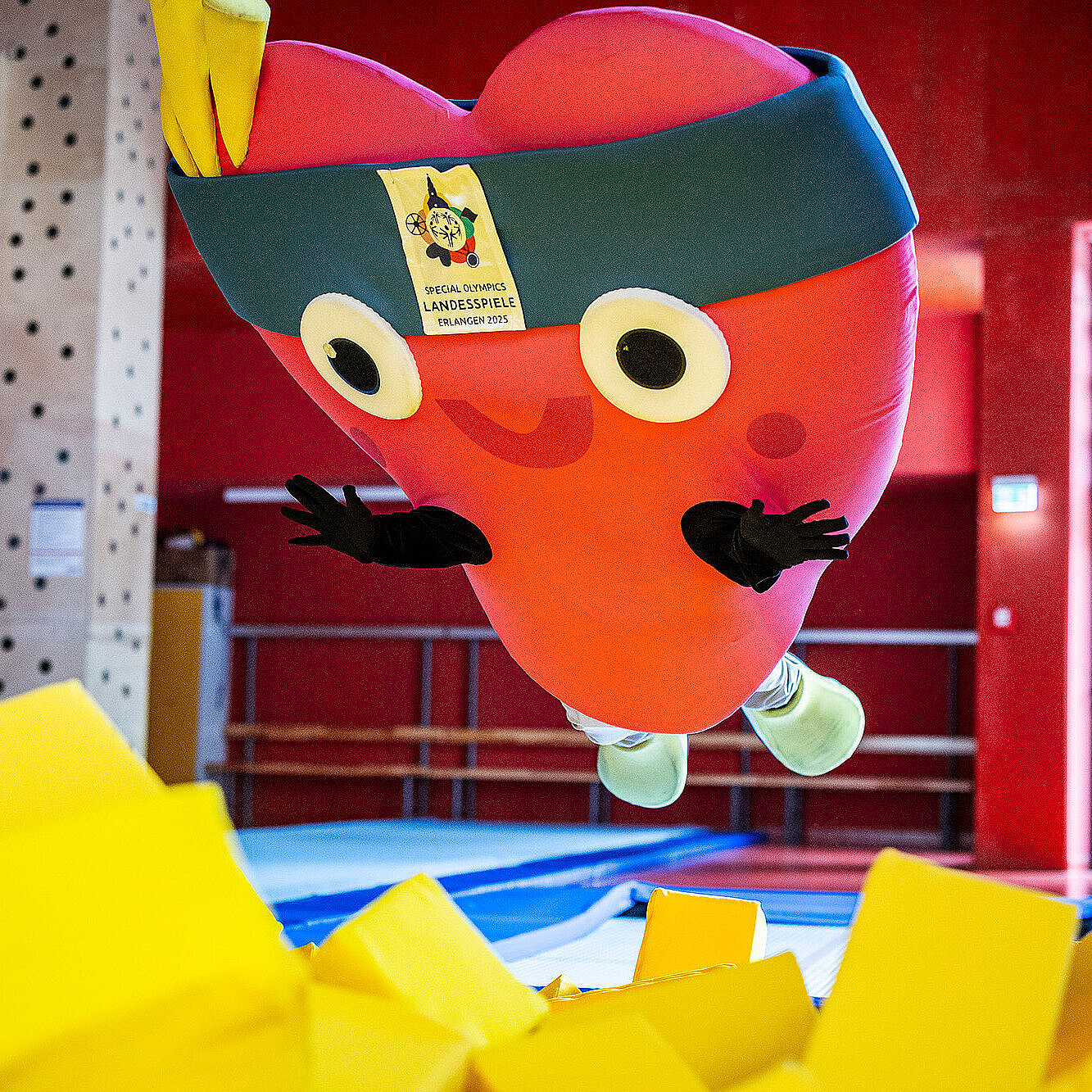 Ein großes, rotes Maskottchen in Herzform mit Stirnband und breitem Lächeln springt in eine gelbe Schaumstoffgrube in einer Sporthalle. Auf dem Stirnband ist ein Schild mit dem Logo und der Aufschrift „Special Olympics Landesspiele Erlangen 2025“ zu sehen.