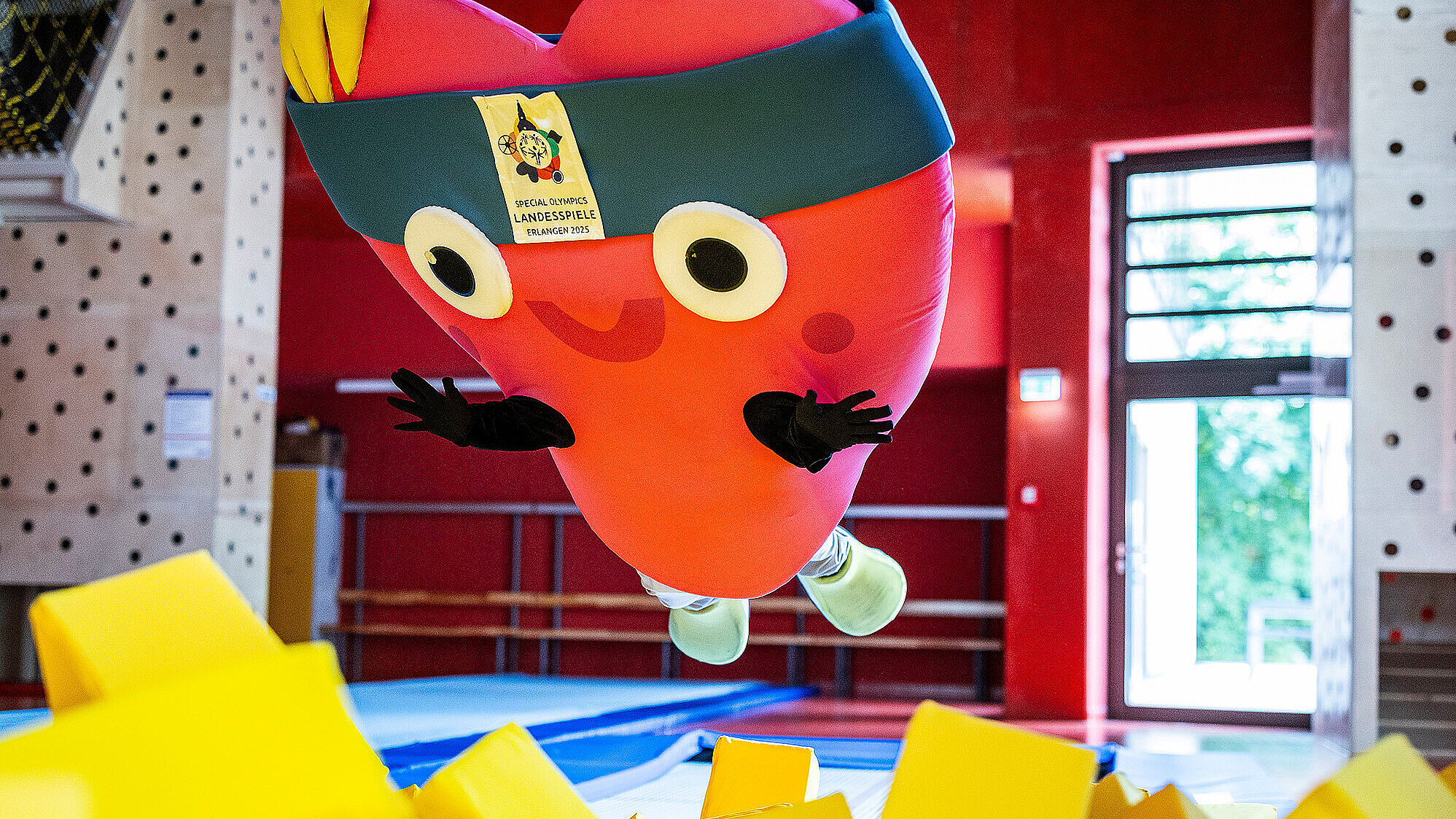 Ein großes, rotes Maskottchen in Herzform mit Stirnband und breitem Lächeln springt in eine gelbe Schaumstoffgrube in einer Sporthalle. Auf dem Stirnband ist ein Schild mit dem Logo und der Aufschrift „Special Olympics Landesspiele Erlangen 2025“ zu sehen.