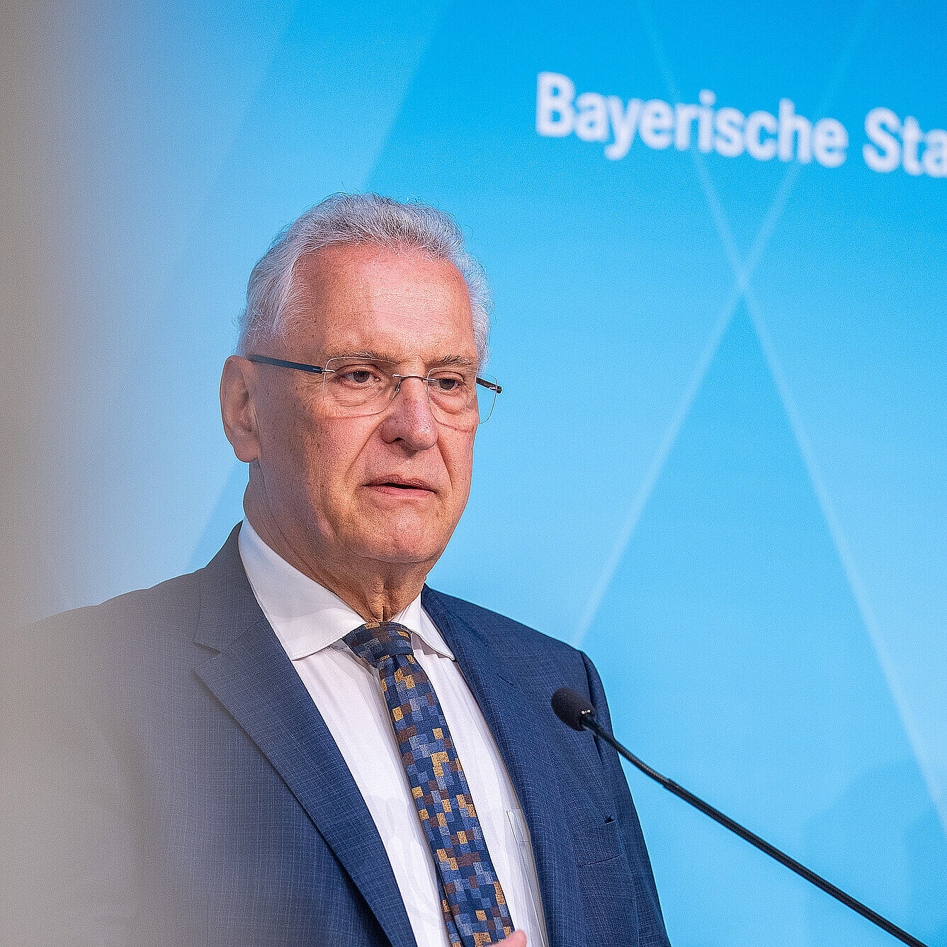 Innenminister Joachim Herrmann steht vor einer blauen Pressewand mit dem Logo Bayerische Staatsregierung an Rednerpulten mit Blick nach vorn.