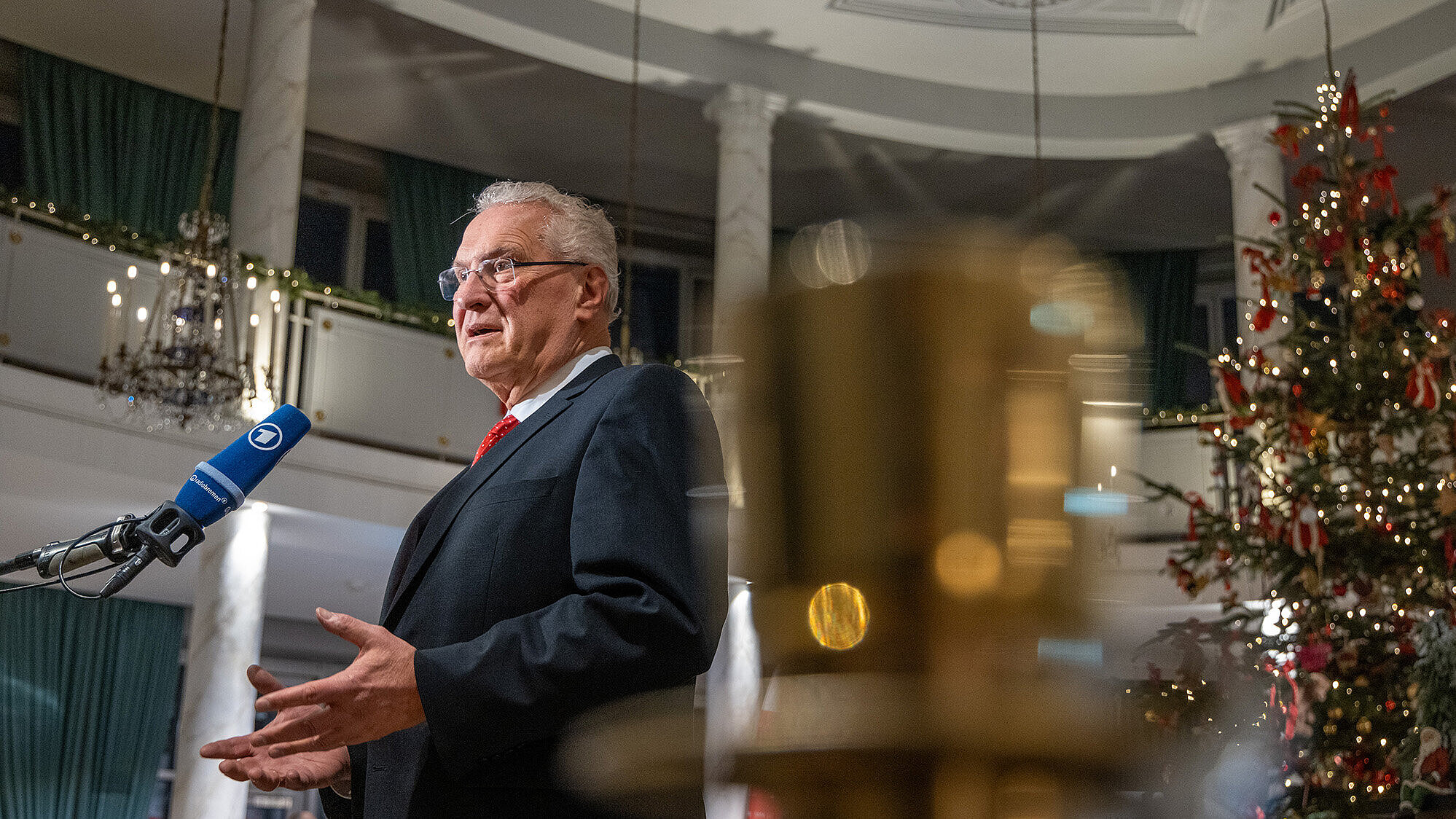 Innenminister Joachim Herrmann in einem dunklen Anzug mit roter Krawatte steht vor einem Mikrofon mit dem Logo der ARD und hält eine Rede oder Ansprache in einem festlich geschmückten Raum mit weißen Marmorsäulen, grünen Vorhängen und einem großen Weihnachtsbaum im Hintergrund. Im Vordergrund ist unscharf ein goldener Kerzenhalter zu sehen.