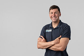 Philipp Nawrath vor hellem Hintergrund, trägt dunkles Poloshirt mit rechteckigem Aufnäher und hat die Arme verschränkt.