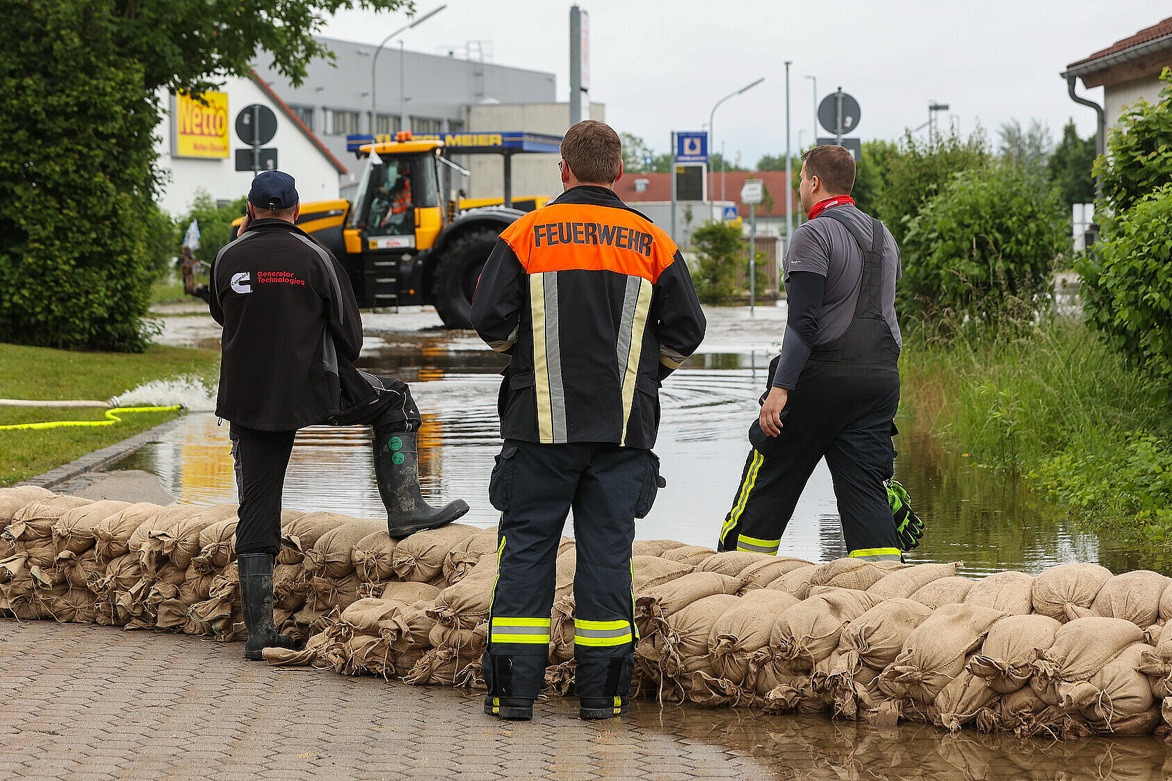 Ein Feuerwehrmann und Hilfskräfte stehen bei Sandsäcken und blicken auf eine überflutete Straße