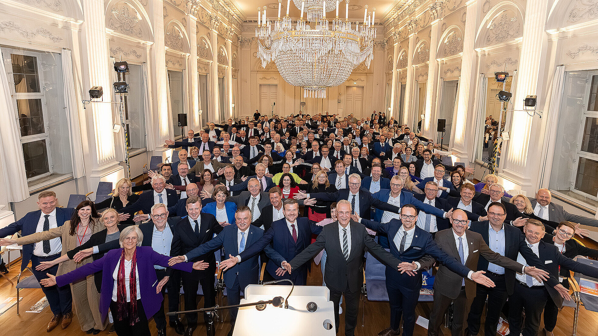 Eine große Gruppe von Menschen steht in einem eleganten Saal mit weißen Wänden und hohen Fenstern. Sie sind in Anzügen und formeller Kleidung gekleidet und haben die Arme ausgestreckt, als würden sie eine Umarmung oder Begrüßung zeigen. Im Vordergrund steht eine Person an einem Rednerpult. Der Saal ist festlich mit einem großen Kronleuchter an der Decke geschmückt.