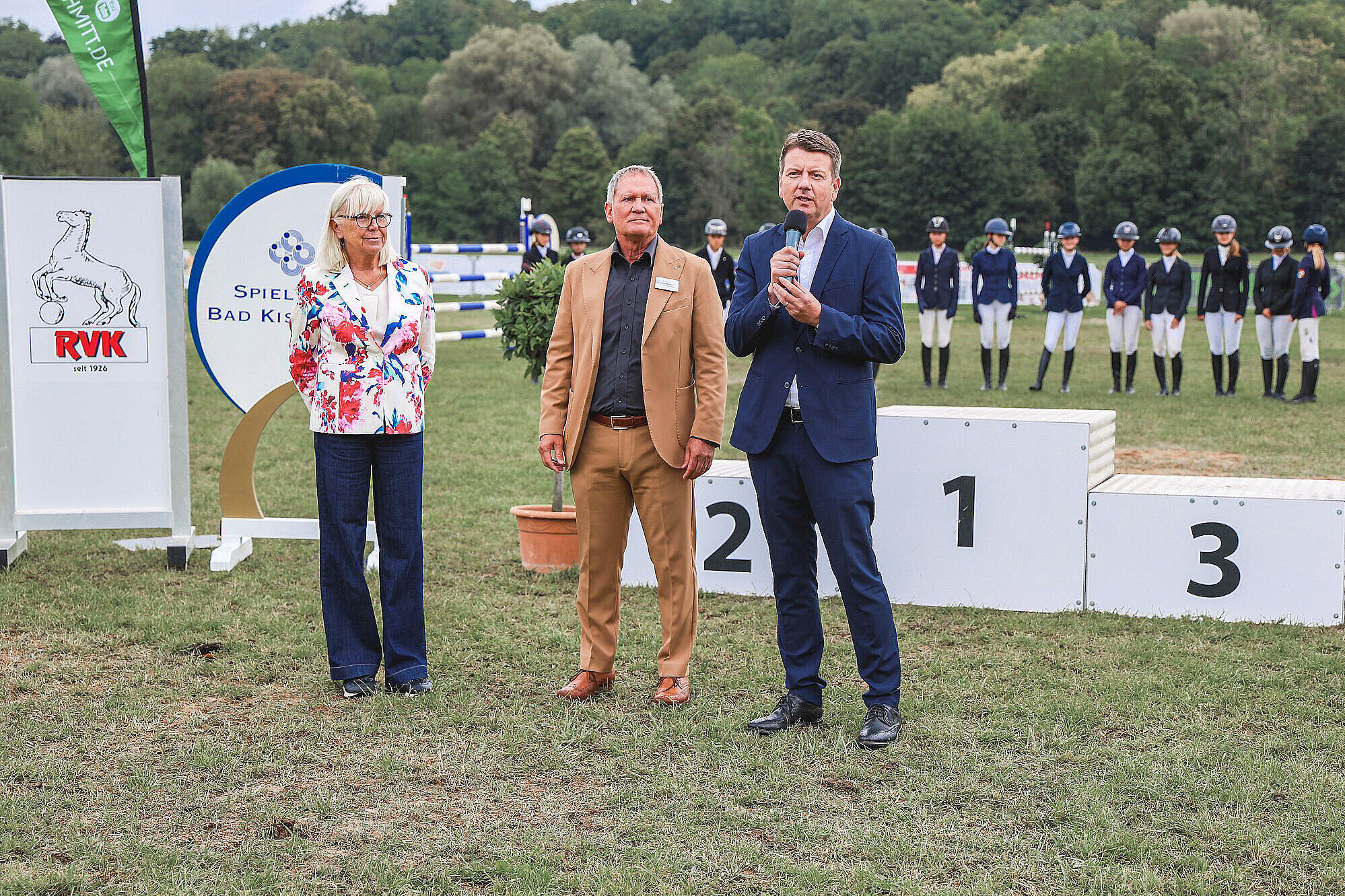 Das Bild zeigt eine Siegerehrung bei einem Reitturnier im Freien. Innenstaatssekretär Sandro Kirchner (re.) und zwei Personen stehen auf dem Rasen vor einem Siegerpodest mit den Platzierungen 1, 2 und 3. Im Hintergrund sind Reiter in Reitkleidung und Helmen in einer Formation aufgestellt. Es ist eine ländliche Umgebung mit Bäumen im Hintergrund.