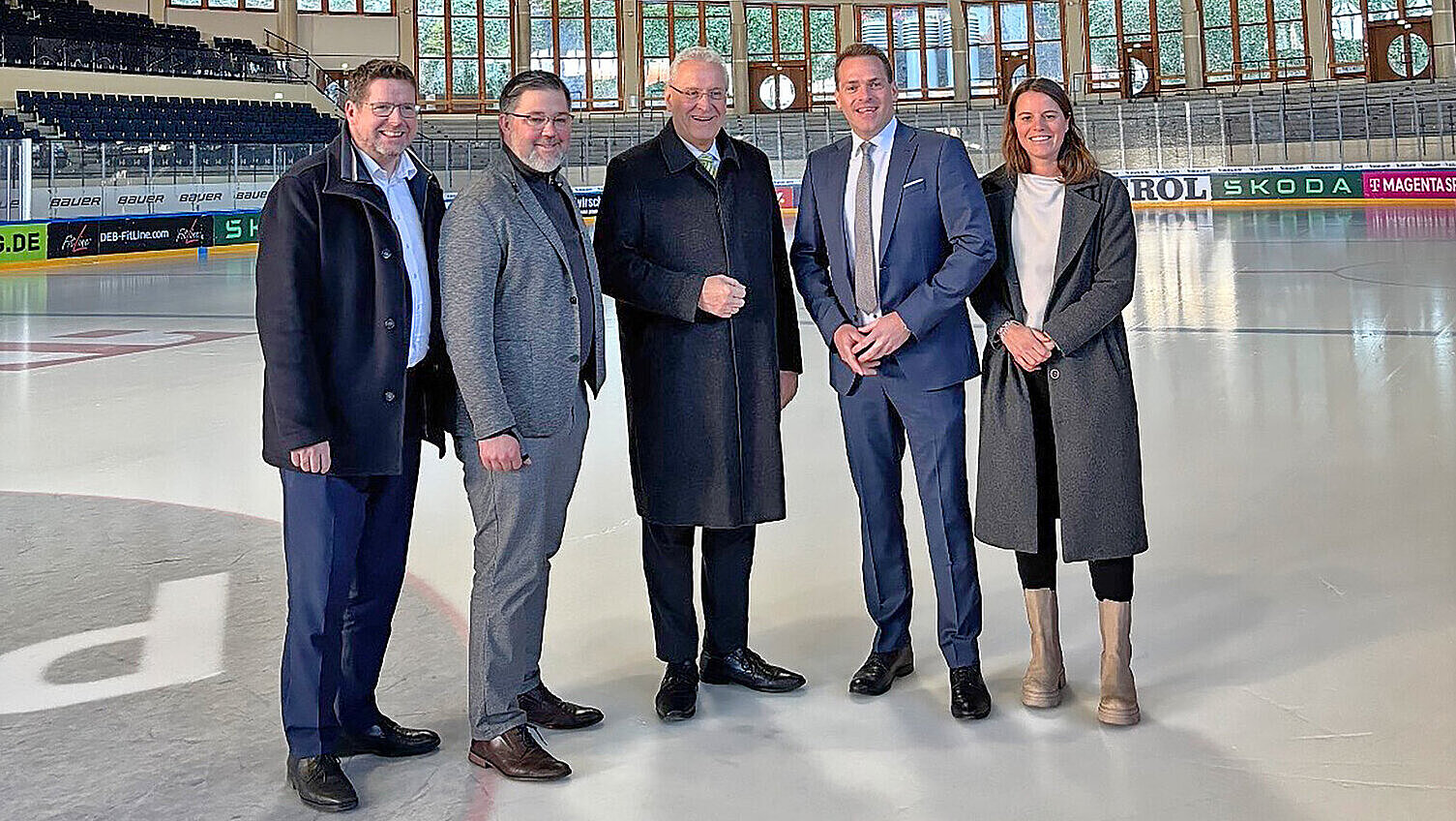 Fünf Personen stehen in einem Eisstadion auf der Eisfläche. Die Halle hat eine markante Holzdecke mit einer großen, hängenden Anzeigetafel in der Mitte. Im Hintergrund sind mehrere Nationalflaggen verschiedener Länder an der Wand angebracht. Die Personen tragen überwiegend formelle Kleidung und stehen in einer Reihe, die Hände teils vor dem Körper gefaltet.