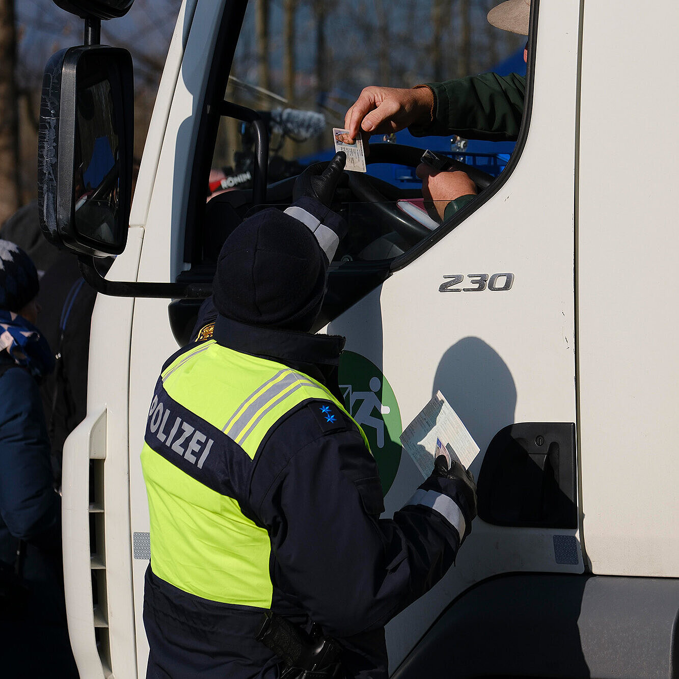Eine Polizistin in einer leuchtend gelben Warnjacke mit der Aufschrift „POLIZEI“ auf dem Rücken hält eine Halte-Kelle mit der Aufschrift „HALT POLIZEI“ in der rechten Hand und stoppt einen weißen Lkw auf einer Straße. Der Lkw ist frontal zu sehen, die Szene spielt offenbar bei Tageslicht.