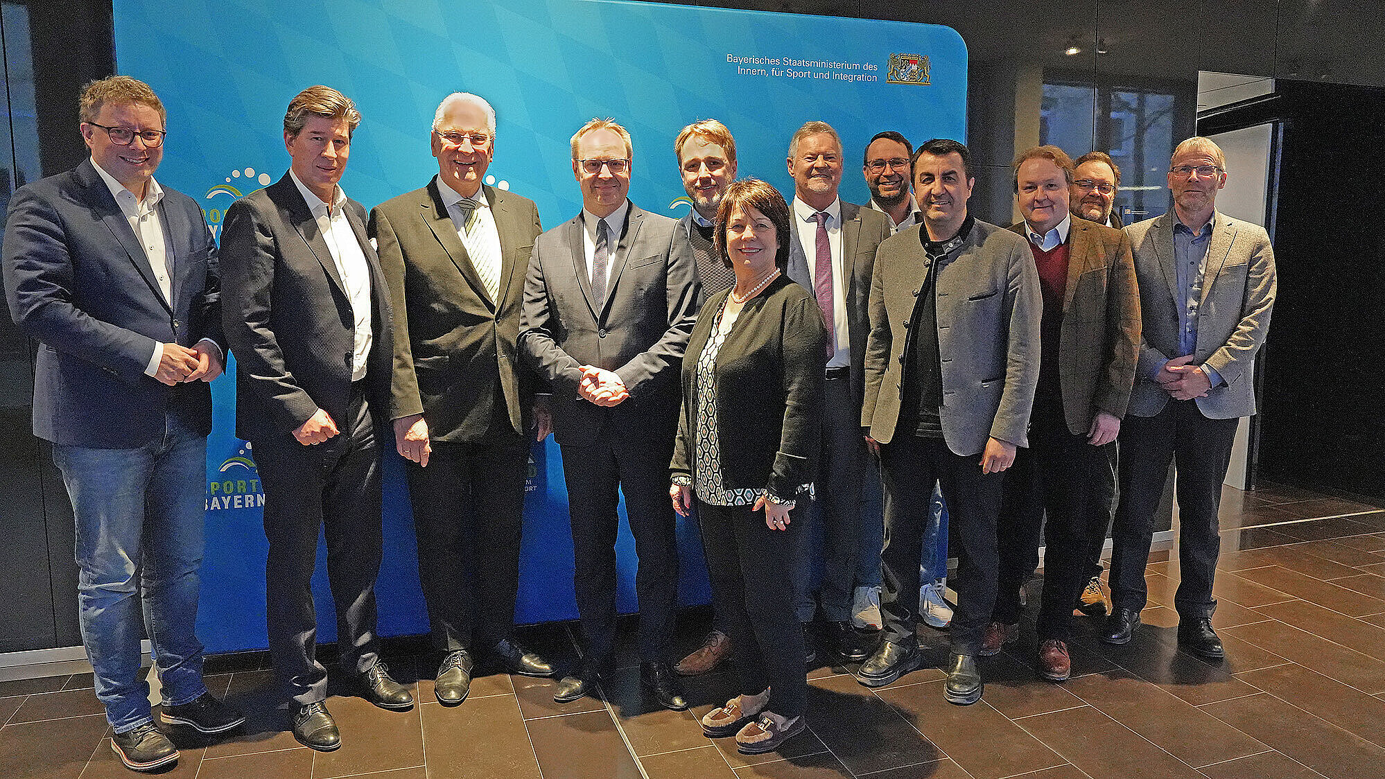 Rund zwölf Personen stehen in zwei Reihen in einem Innenraum vor einem blauen Messestand mit bayerischem Logo; die Personen tragen überwiegend Anzüge und Jacken.