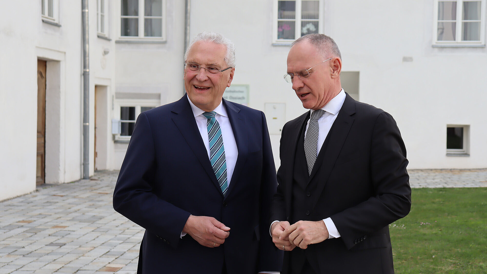 Zwei Männer in dunklen Anzügen stehen in einem gepflasterten Innenhof vor einem weißen Gebäude; Gesichter unkenntlich gemacht.