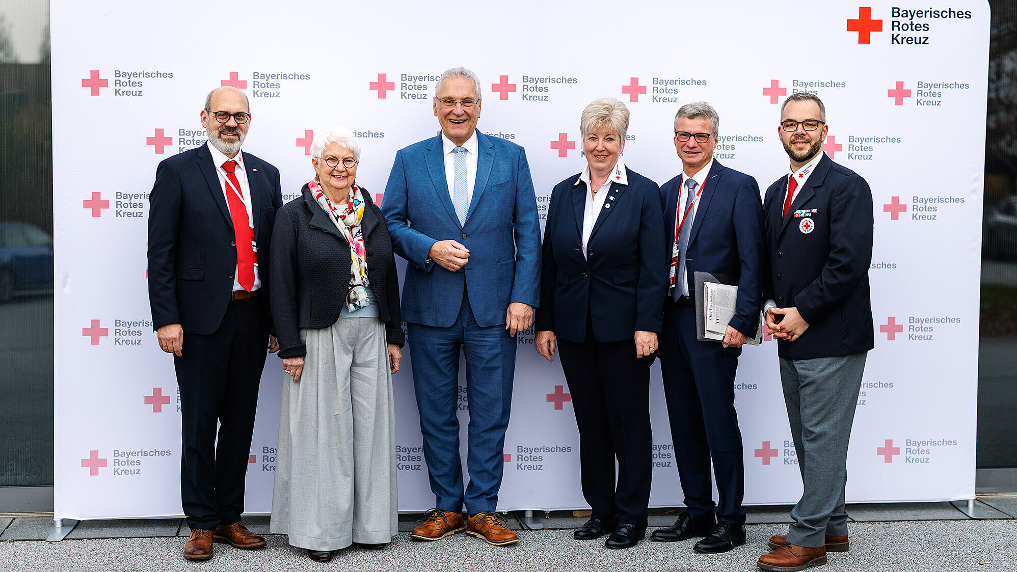 Sieben Personen stehen vor einer Fotowand (3. von li. Innenminister Joachim Herrmann) mit dem Logo des Bayerischen Roten Kreuzes. Alle sind formell gekleidet, sechs Männer tragen Anzüge, zwei davon mit roten Krawatten, und eine Frau trägt einen dunklen Blazer mit weißer Bluse. Eine Person hält eine Mappe in der Hand. Der Hintergrund ist hell und zeigt mehrfach das Logo des Bayerischen Roten Kreuzes.