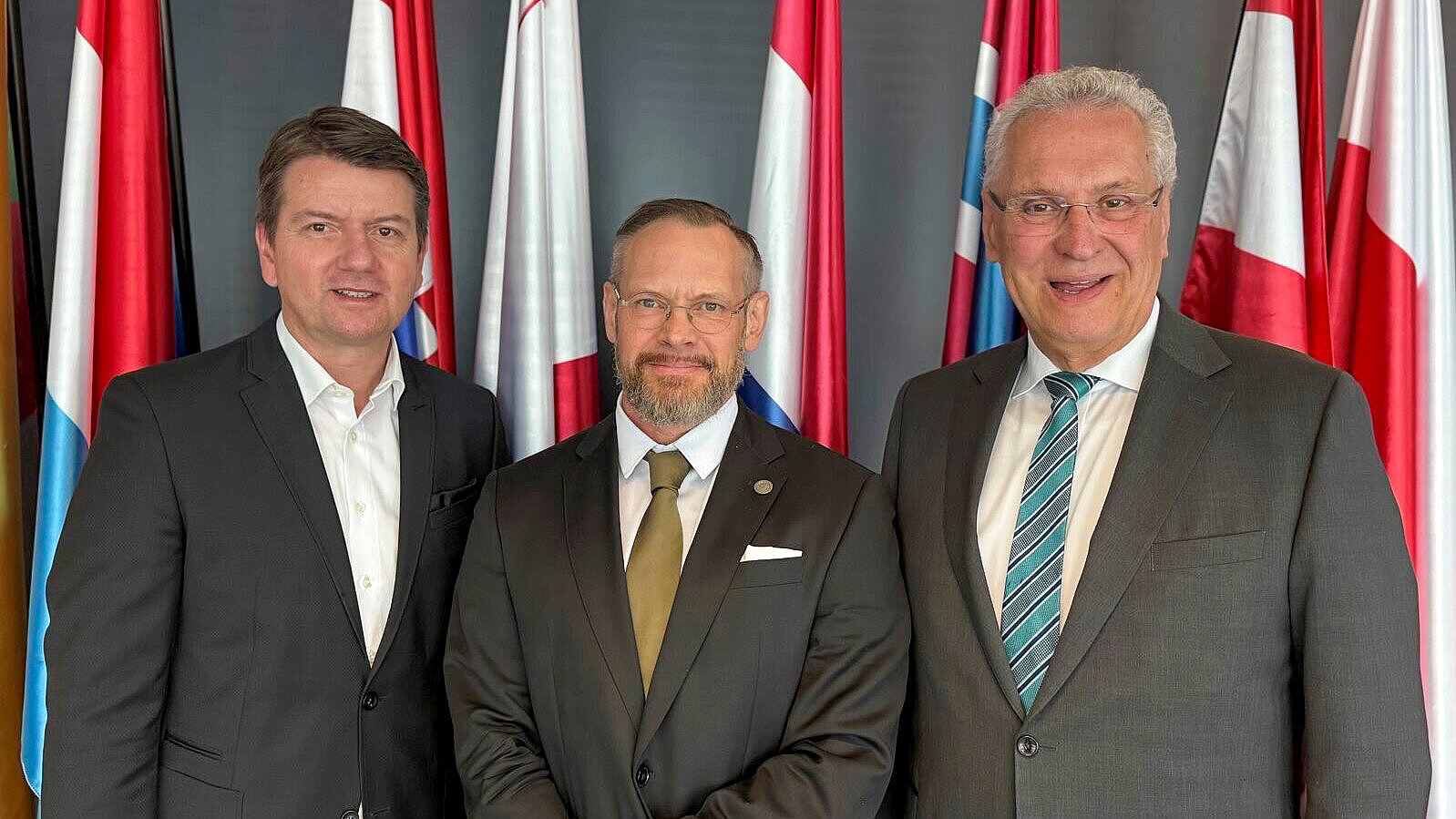 links Innenstaatssekretär Sandro Kirchner, rechts Innenminister Joachim Herrmann mit Blick in die Kamera vor einem Fahnenset