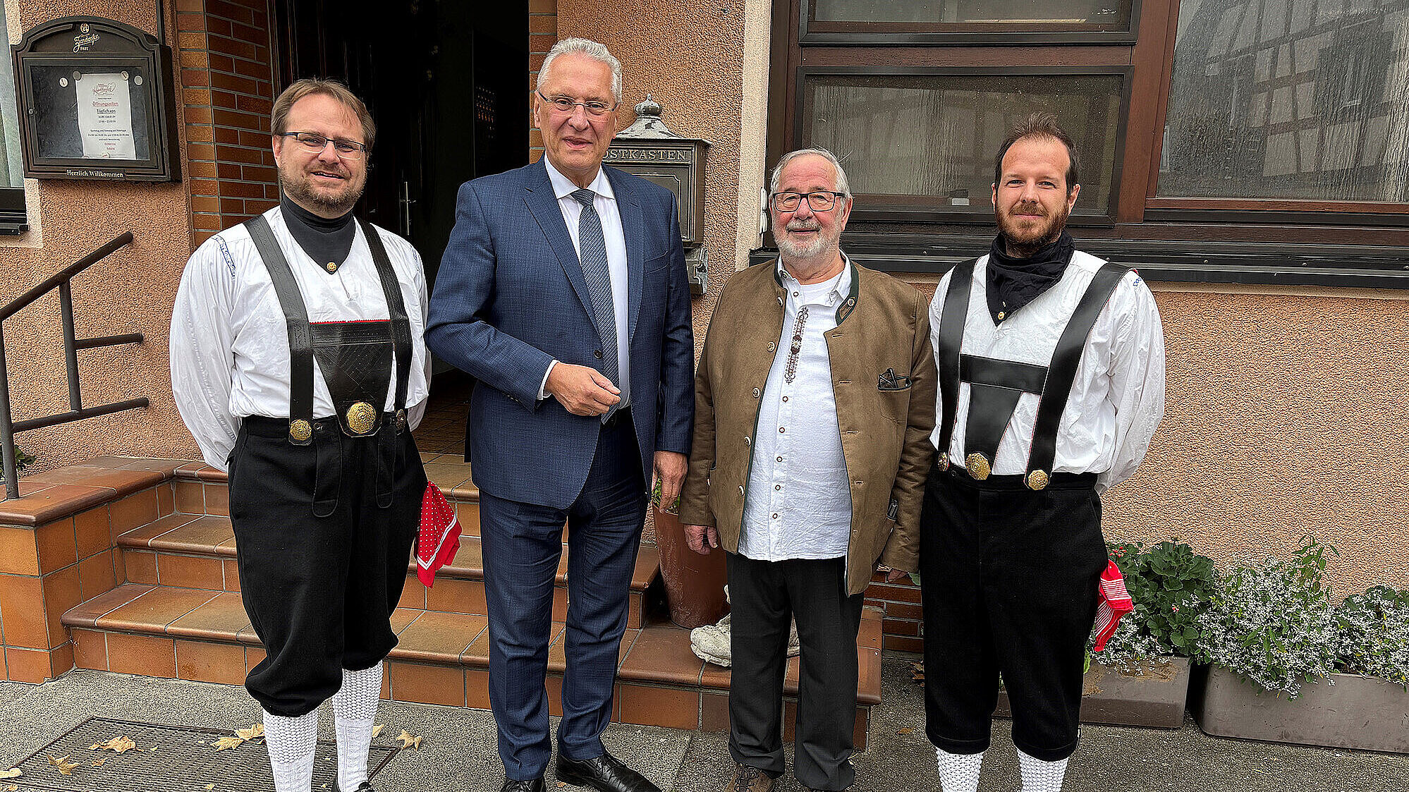 Vier Männer stehen nebeneinander vor einem Gebäude (2.v.li. Innenminister Joachim Herrmann). Zwei von ihnen tragen traditionelle Trachten mit schwarzen Hosen, weißen Hemden, schwarzen Gürteln mit goldenen Schnallen, weißen Strümpfen und schwarzen Schuhen. Der Mann in der Mitte trägt einen dunklen Anzug mit Krawatte. Der zweite Mann von rechts trägt eine beige Jacke über einem weißen Hemd. Das Gebäude im Hintergrund hat eine braune Fassade, eine Treppe und Fenster. Es liegen einige Blätter auf dem Boden.
