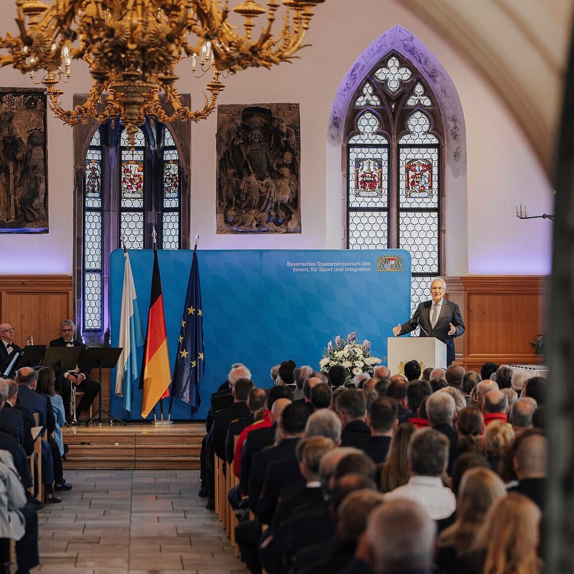 Das Bild zeigt den Festsaal, man sieht in zwei Reihen das Publikum sitzen und auf einer Bühne Innenminister Joachim Herrmann zu den Anwesenden sprechen. Auf einer Leinwand steht der Name der Veranstaltung.
