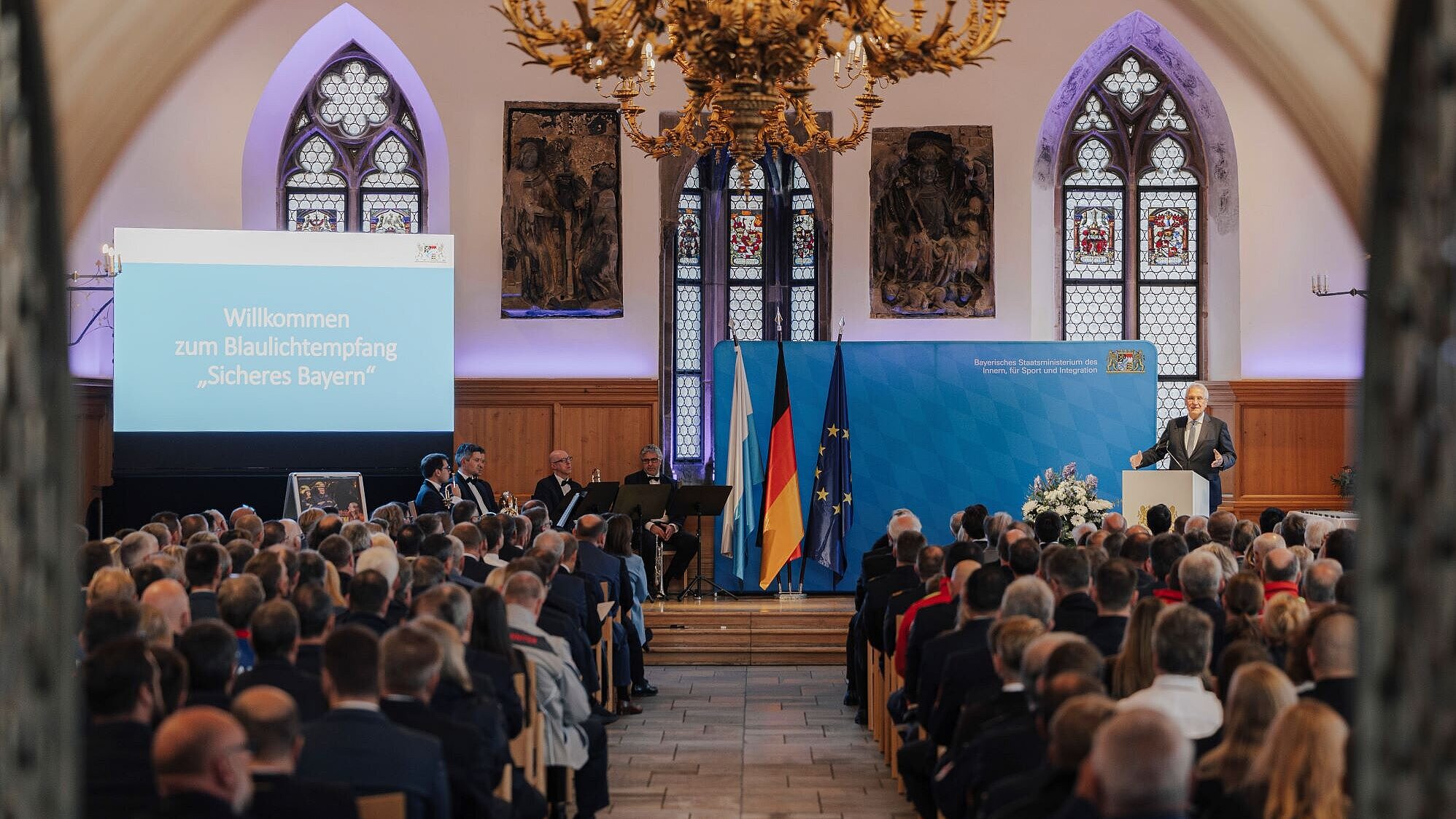 Das Bild zeigt den Festsaal, man sieht in zwei Reihen das Publikum sitzen und auf einer Bühne Innenminister Joachim Herrmann zu den Anwesenden sprechen. Auf einer Leinwand steht der Name der Veranstaltung.