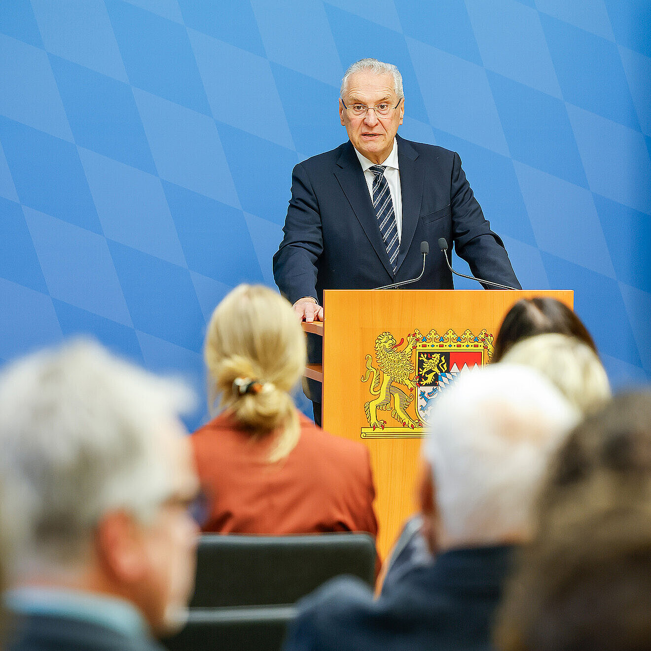 Innenminister Joachim Herrmann im dunklem Anzug und gestreifter Krawatte steht hinter einem Rednerpult mit dem bayerischen Wappen und hält eine Rede. Im Vordergrund sitzen mehrere Personen, die dem Vortrag zuhören. Der Hintergrund zeigt ein blaues Muster mit Rauten.