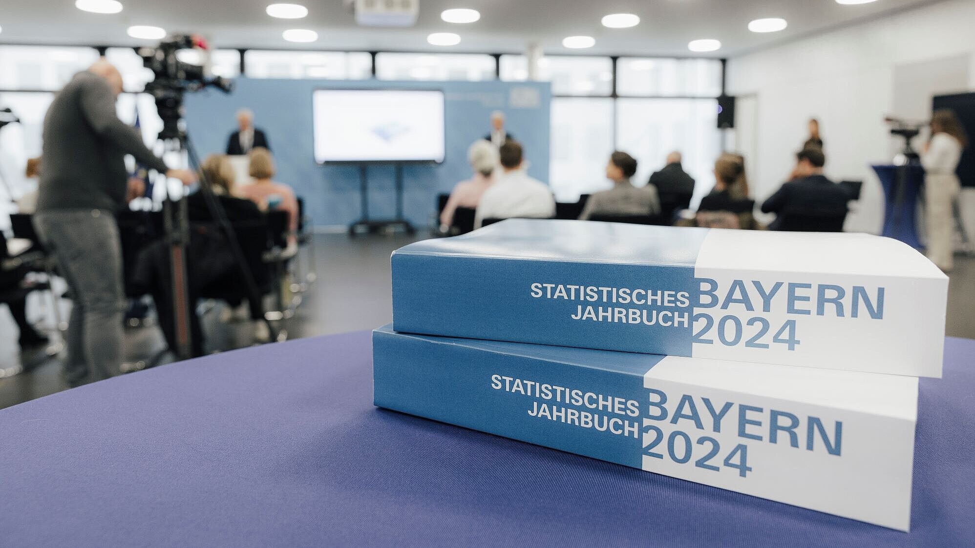 Auf dem Bild sind im Vordergrund zwei Exemplare des Statistischen Jahrbuchs für Bayern 2024 in Buchform zu sehen, im Hintergrund die laufende Pressekonferenz zur Vorstellung.