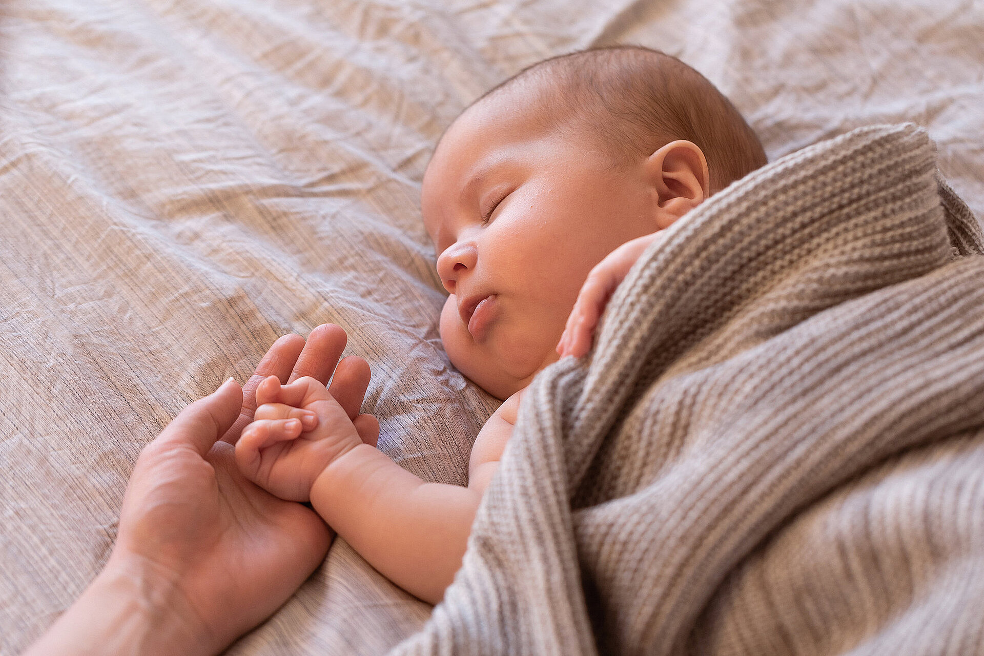 Eine Mutter hält die Hand eines schlafenden Babys, das mit einer Decke zugedeckt ist