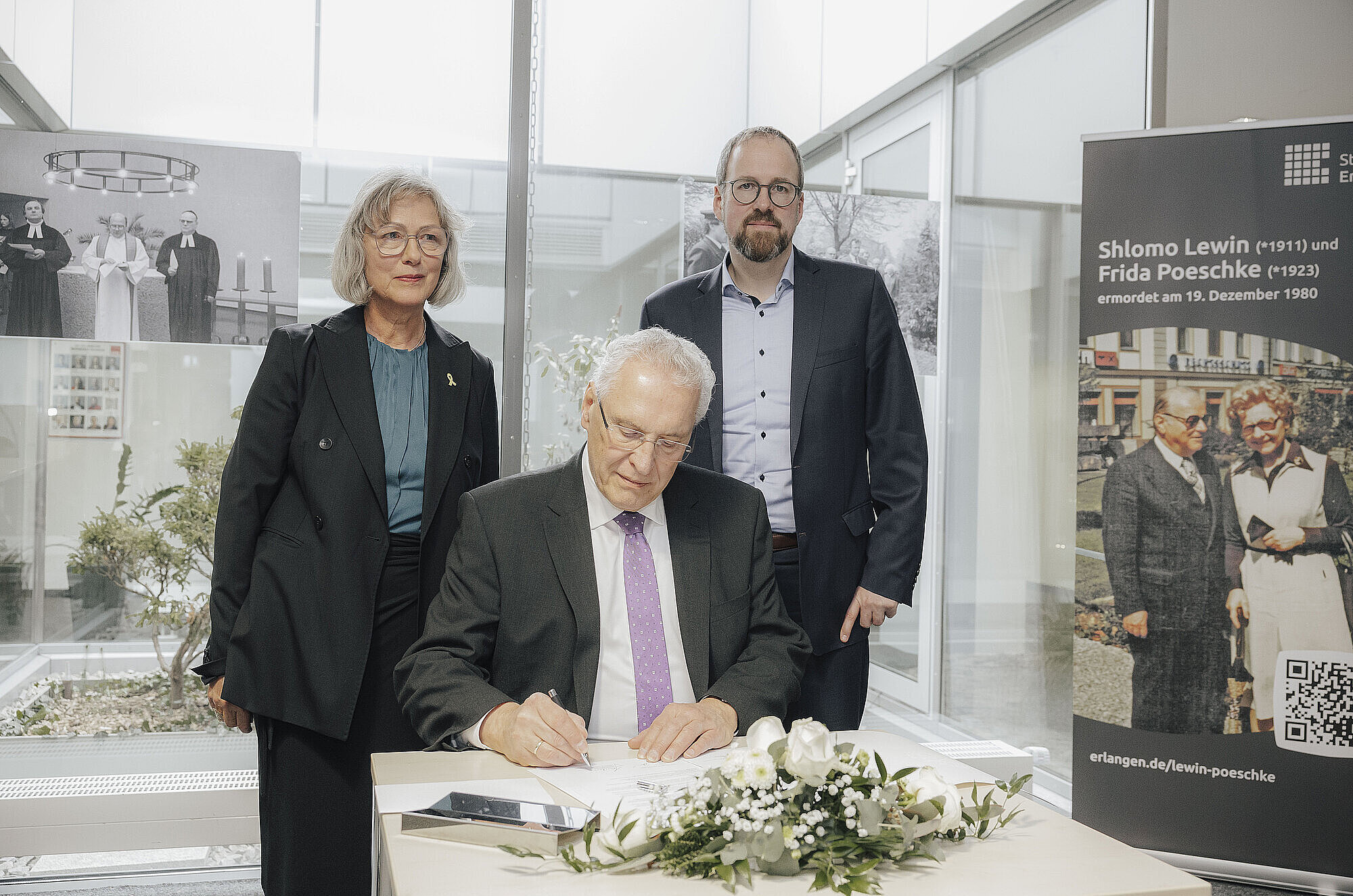 Auf dem Bild ist Innenminister Joachim Herrmann zu sehen, wie er sich in ein Gedenkbuch anlässlich der Veranstaltung einträgt. Hinter ihm stehen zwei Personen und blicken in die Kamera.