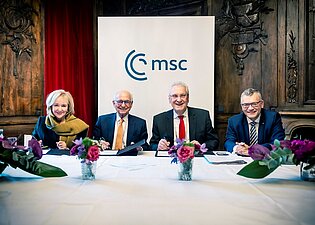 Vier Personen sitzen an einem Tisch, hinter ihnen ein Banner mit der Aufschrift "MSC".